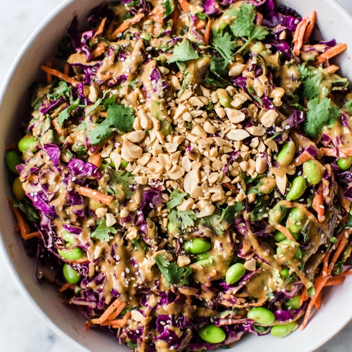 Crunchy Thai Peanut Salad in a white bowl, featuring shredded purple cabbage, carrots, and edamame tossed in a creamy peanut dressing, garnished with chopped peanuts and cilantro.