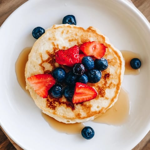 Golden-brown Cottage Cheese Pancakes stacked high on a white plate, served with fresh blueberries and a drizzle of warm maple syrup.
