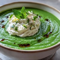 A bowl of vibrant spring pea soup topped with fluffy whipped ricotta and a drizzle of bright green mint oil.